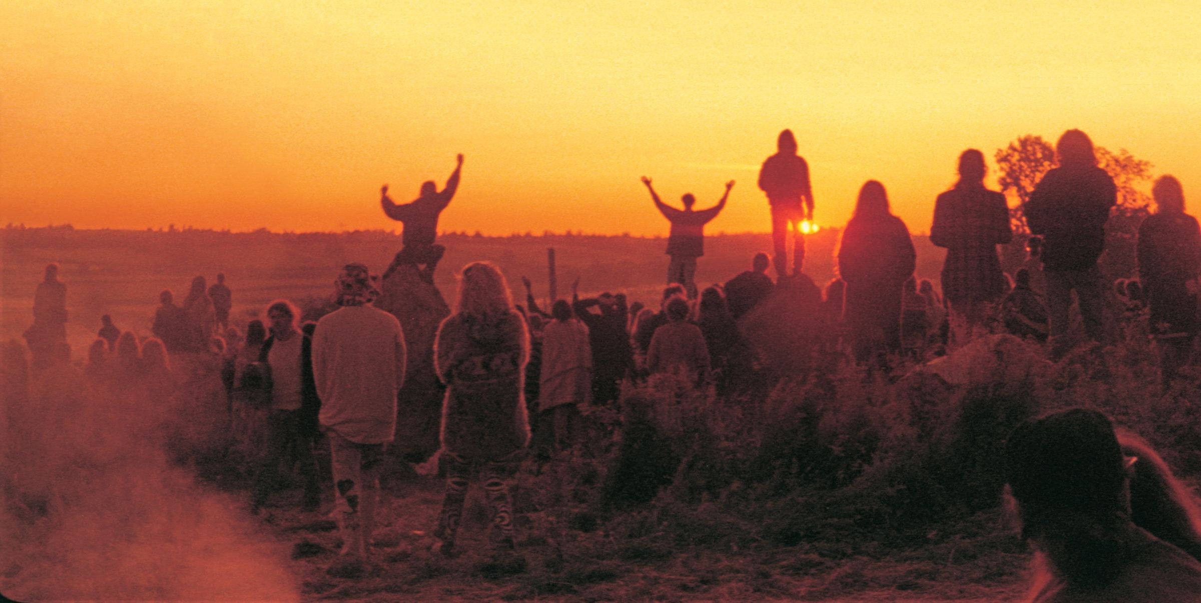 Sunrise over the crowd at Glastonbury in 1993, figures with arms raised against an orange sky
