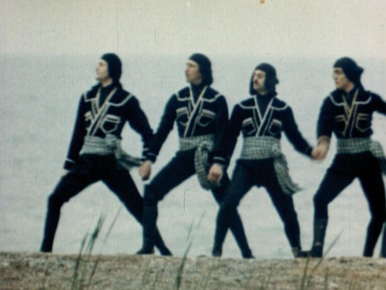 Four performers in black costumes holding hands on a shoreline