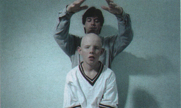 A bald boy in a white football shirt sits still while a man behind him raises his hands over his head